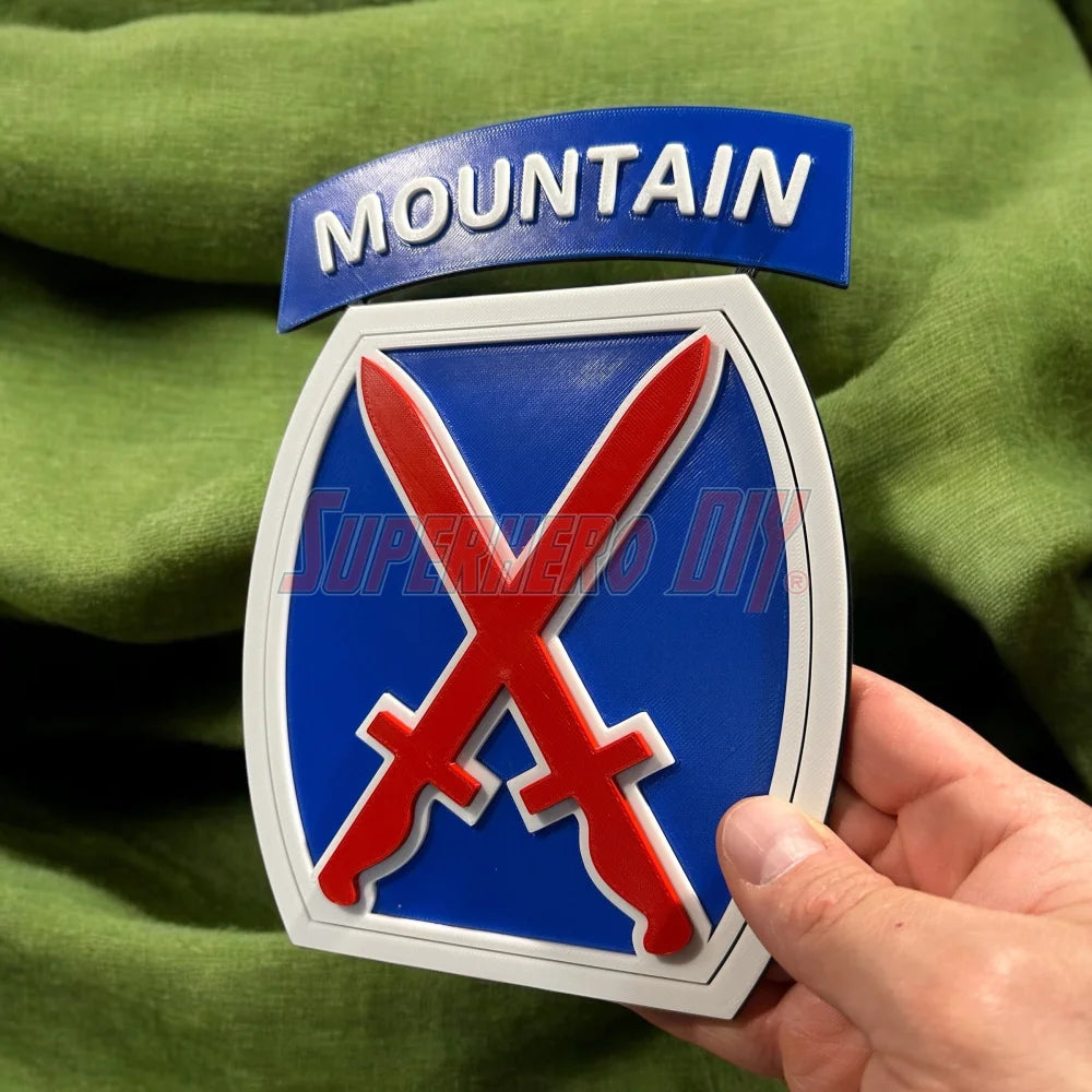 3D-printed 10th Mountain Division insignia in blue, white, and red with crossed swords, held against a green background.