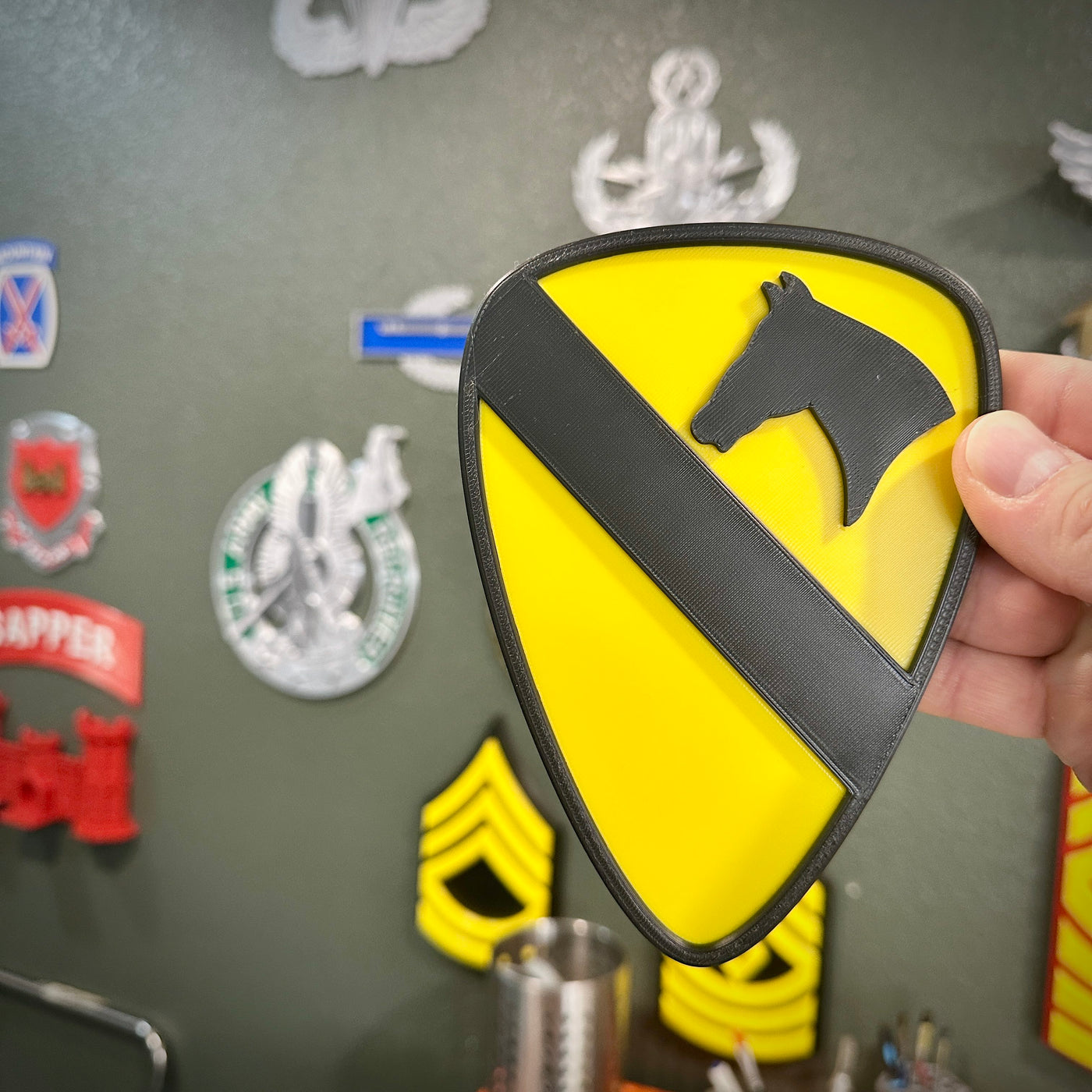 Person holding a 1st Cavalry Division Display Insignia in front of a wall with military decor.