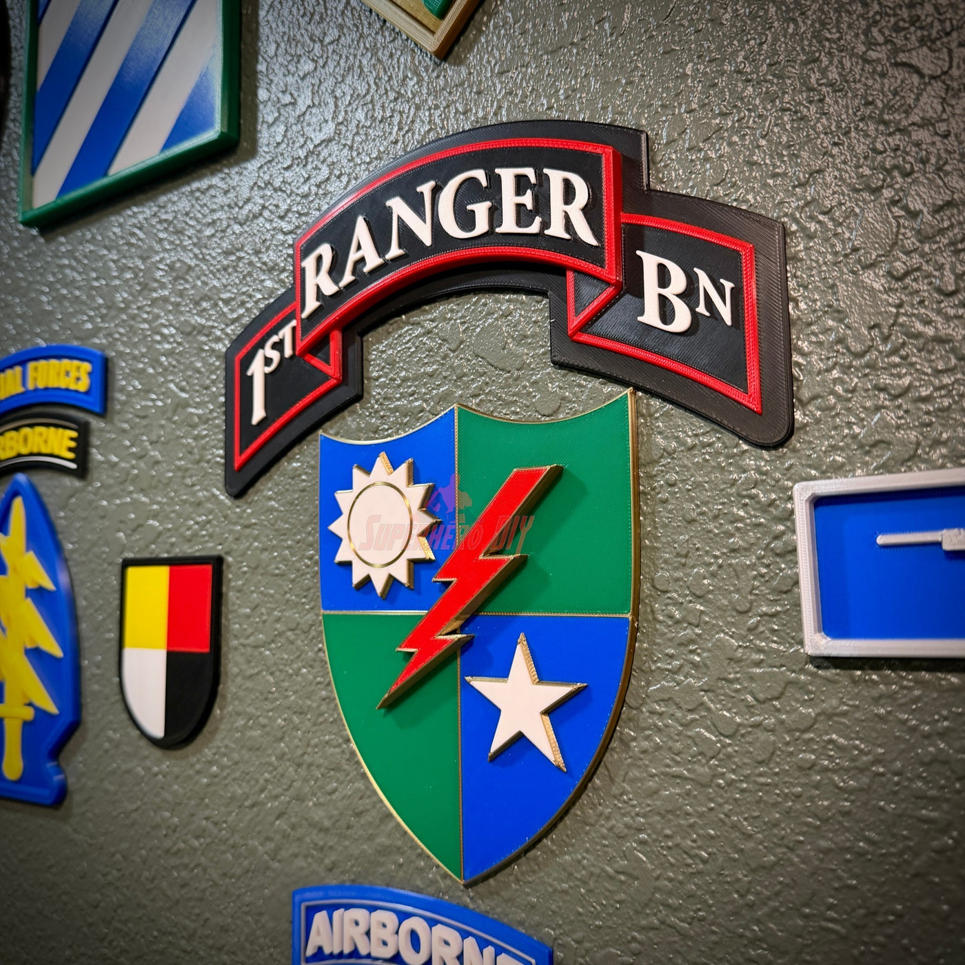 A military-style patch or emblem mounted on a textured gray wall, featuring a black and red banner reading '1st Ranger BN' above a blue and green shield with a white sun, red lightning bolt, and white star.