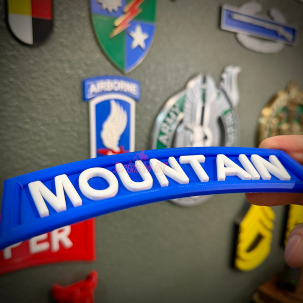 3D mountain tab wall display in blue with raised white lettering being held up against decorative military patches.