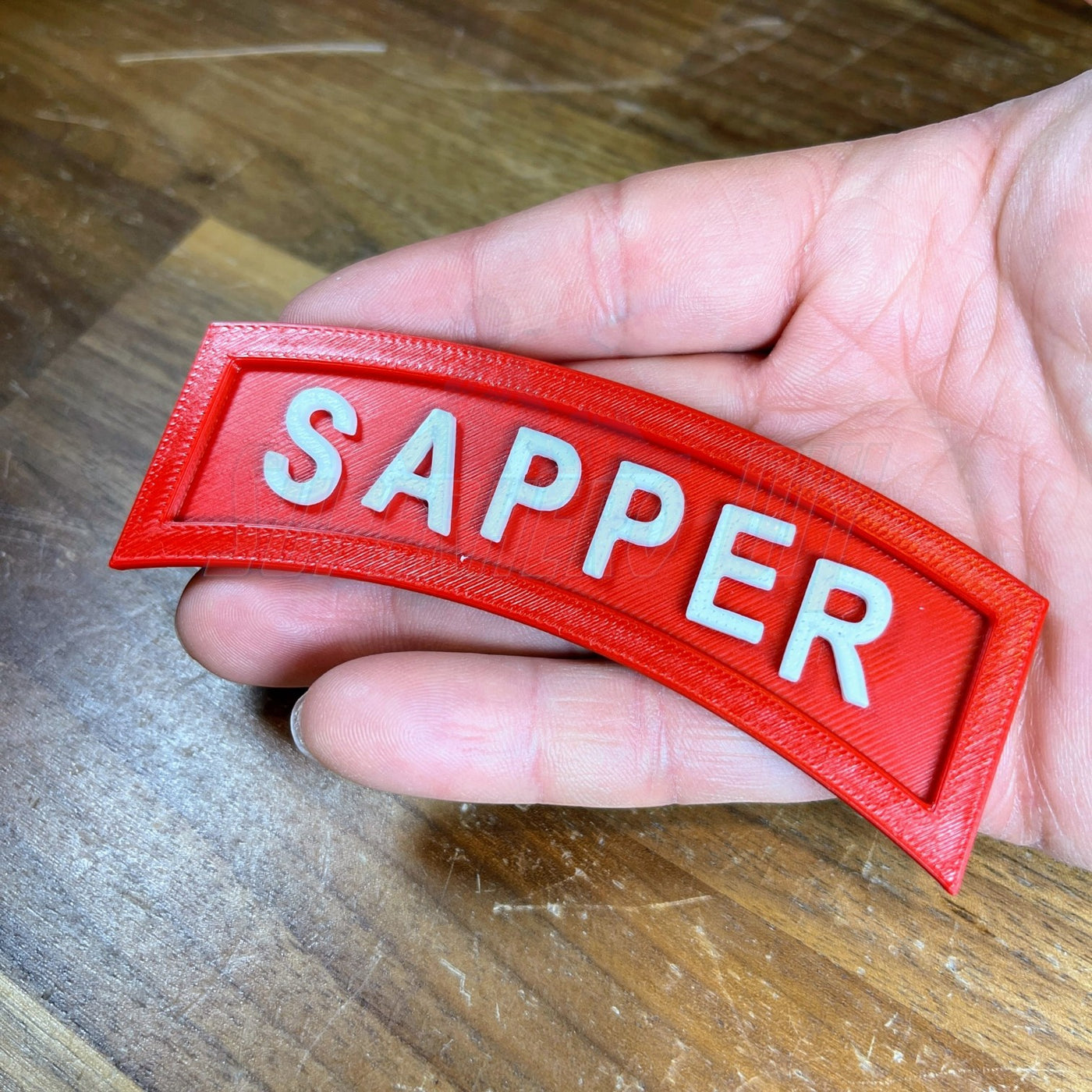 3D sapper tab displayed in a hand, showcasing the bold lettering against a vibrant red background.