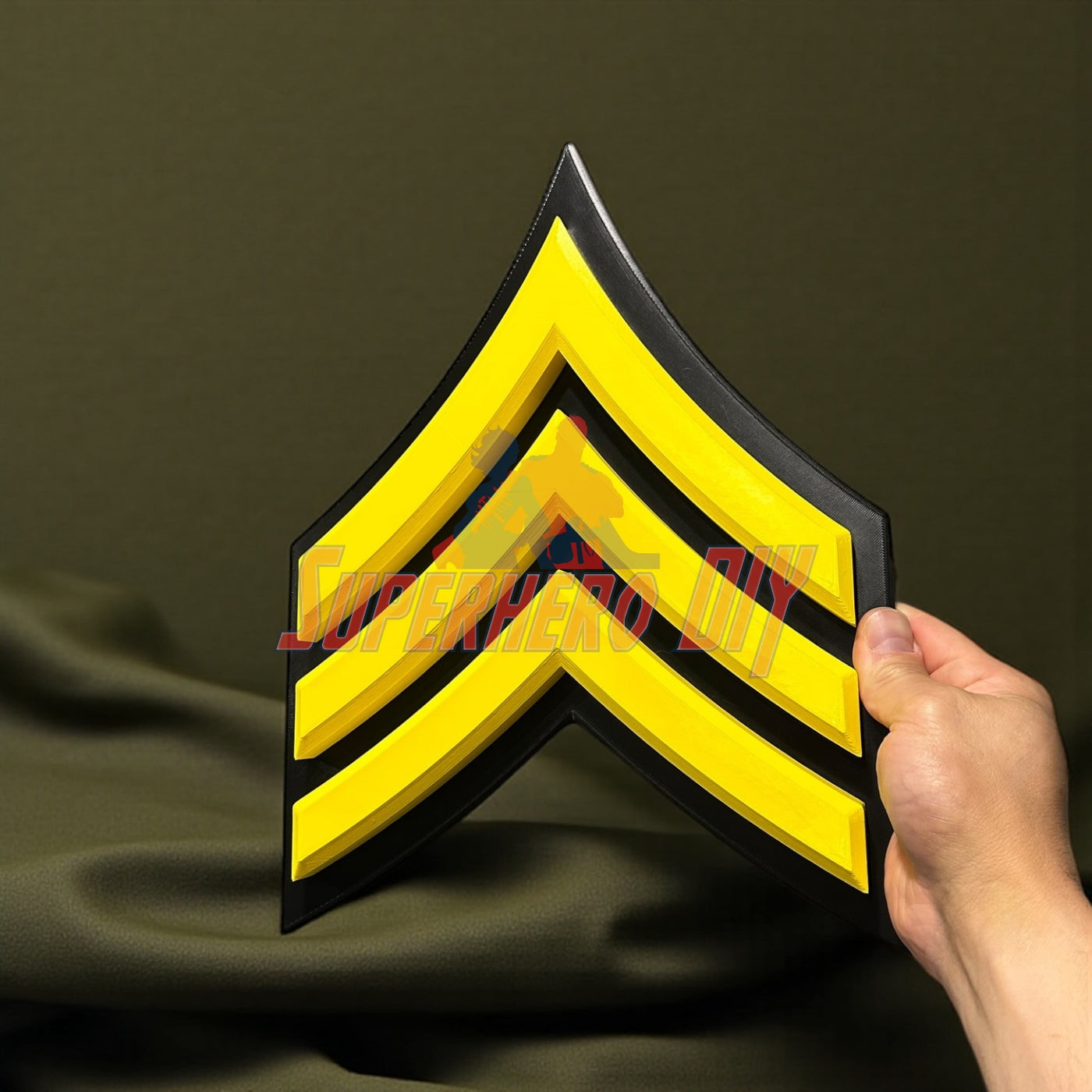Hand holding a bold, 3D-printed Army enlisted rank insignia in yellow and black.