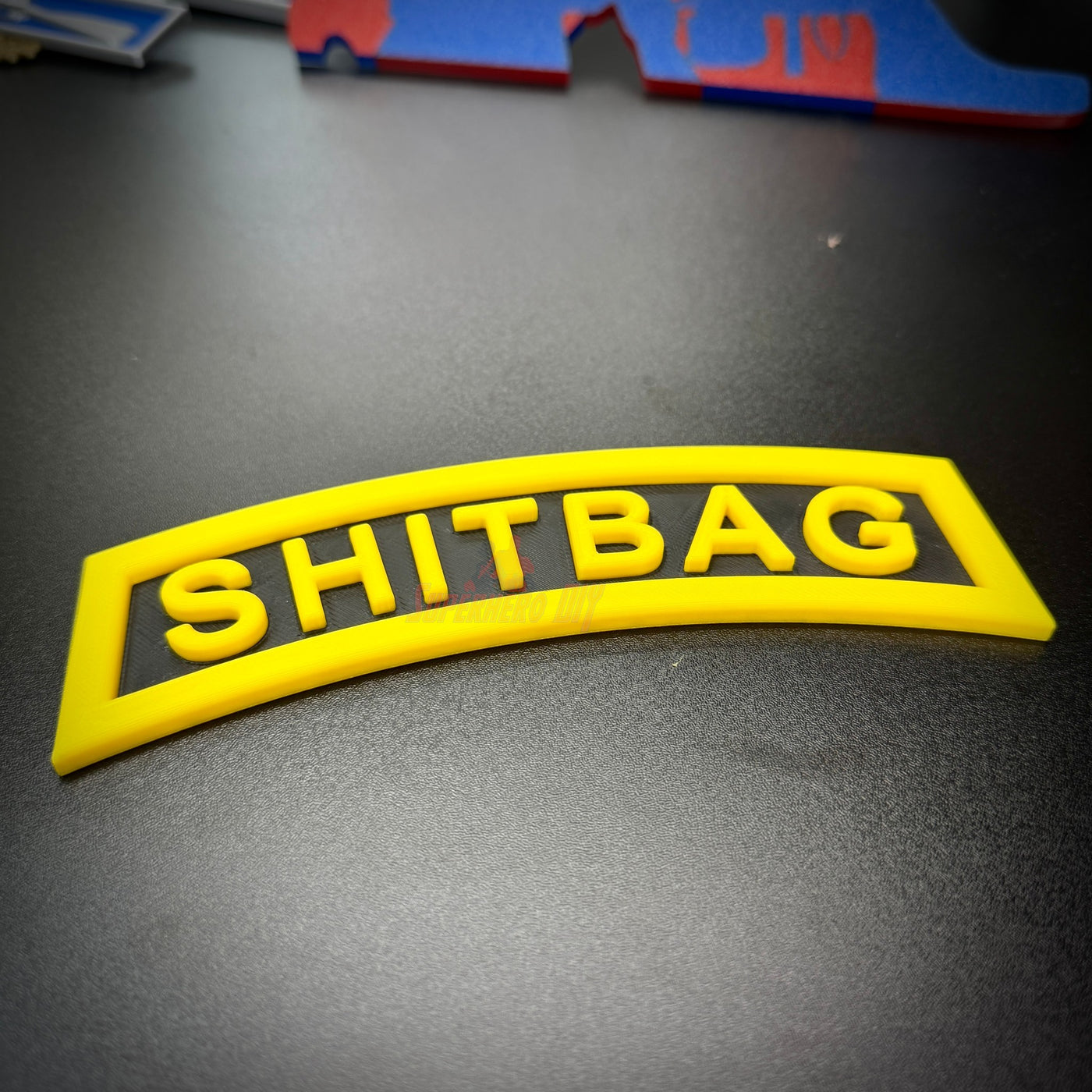SHITBAG tab displayed on a black surface, featuring a classic arched design in yellow and black.
