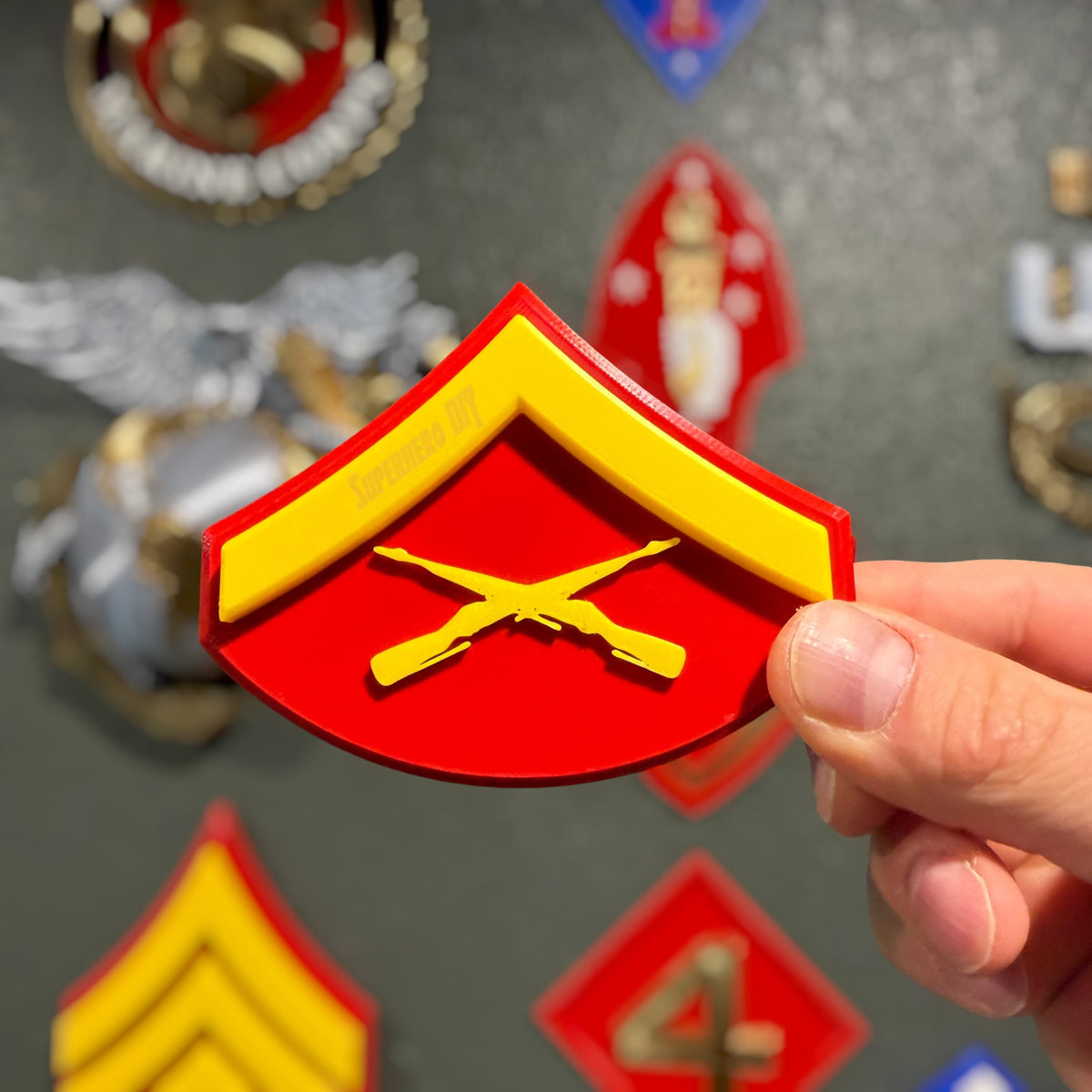 Hand holding a red and yellow USMC Lance Corporal rank insignia, other Marine insignia displays blurred behind