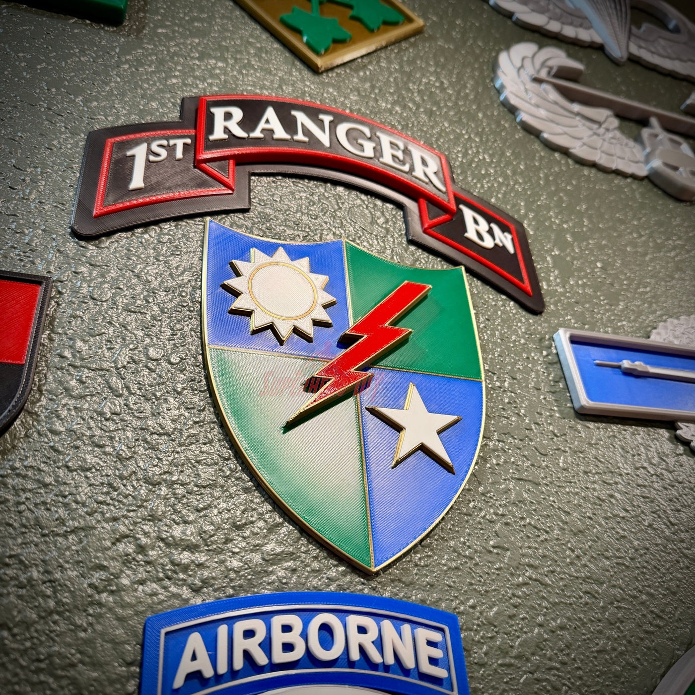 A close-up of military-style enamel patches on a textured gray surface, featuring a central shield patch with blue, green, white, red, and gold sections, plus a red '1ST RANGER BN' ribbon patch above it and an 'AIRBORNE' patch at the bottom.