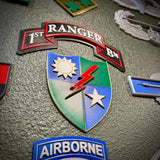 A close-up of military-style enamel patches on a textured gray surface, featuring a central shield patch with blue, green, white, red, and gold sections, plus a red '1ST RANGER BN' ribbon patch above it and an 'AIRBORNE' patch at the bottom.