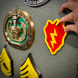 25th Infantry Division insignia being pressed onto a gray wall beside other military insignia and rank chevron displays
