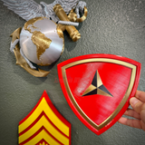 3rd Marine Division insignia held by a hand, featuring a red shield with gold accents and three-pointed caltrap.