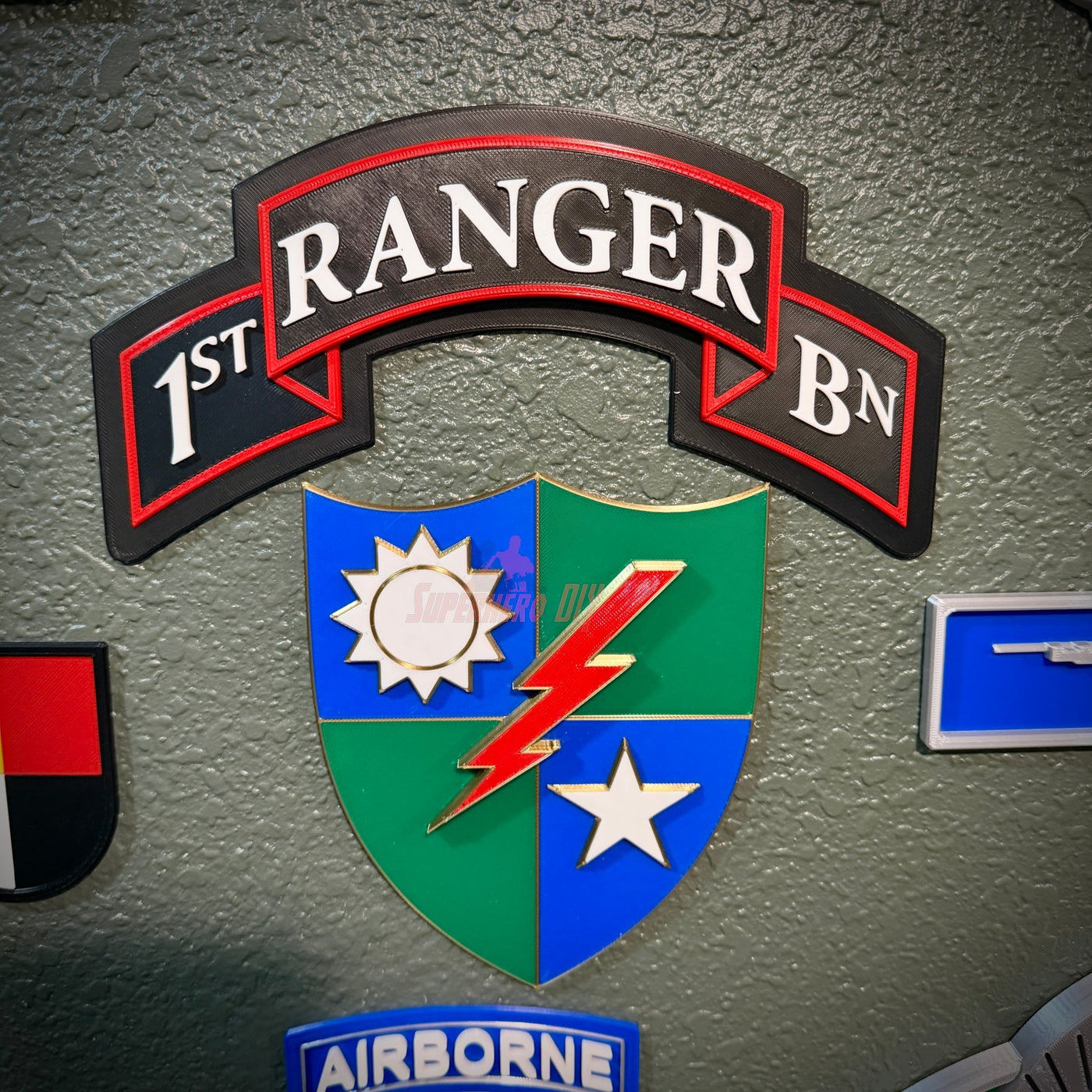 A 1st Ranger Battalion insignia patch mounted on a textured gray wall, featuring a black arched banner with white lettering and a central shield in blue, green, red, and white.