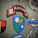 A military patch or badge featuring a shield with blue and green quadrants, a red lightning bolt, a white star, and a white banner above reading "RANGER" with "1ST BN" on either side, mounted on a textured olive-green wall.