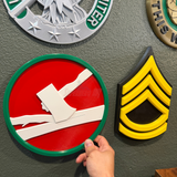 Hand holding the 84th Training Command display insignia plaque on a wall with other military insignias.