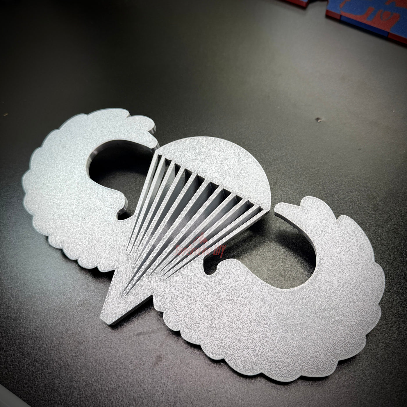 Replica airborne jump wings badge displayed on a black surface, showcasing the iconic wings and parachute design.