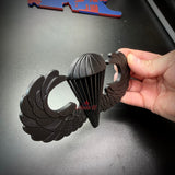 Hand holding an airborne jump wings badge with black finish and detailed wing design.