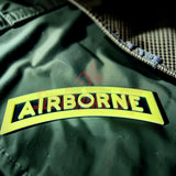 3D airborne tab displayed on a green fabric, featuring bold yellow lettering, showcasing military insignia.