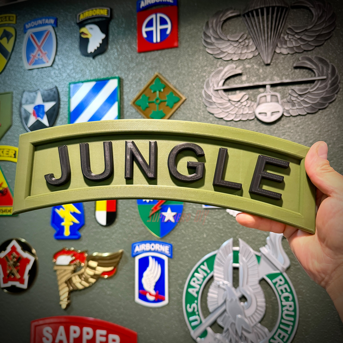 A hand holding a 3D jungle tab display piece with black lettering on an army green background.