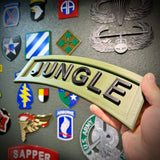 A hand holding a 3D jungle tab display piece in army green with black lettering against a wall of military insignias.