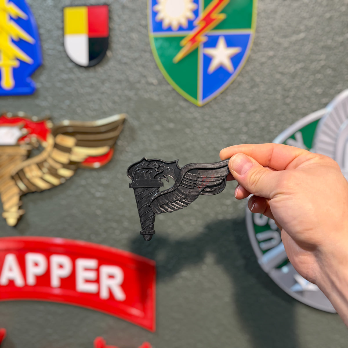 A hand holds a black Pathfinder badge emblem against a gray wall with other military-style badges and patches in the background.
