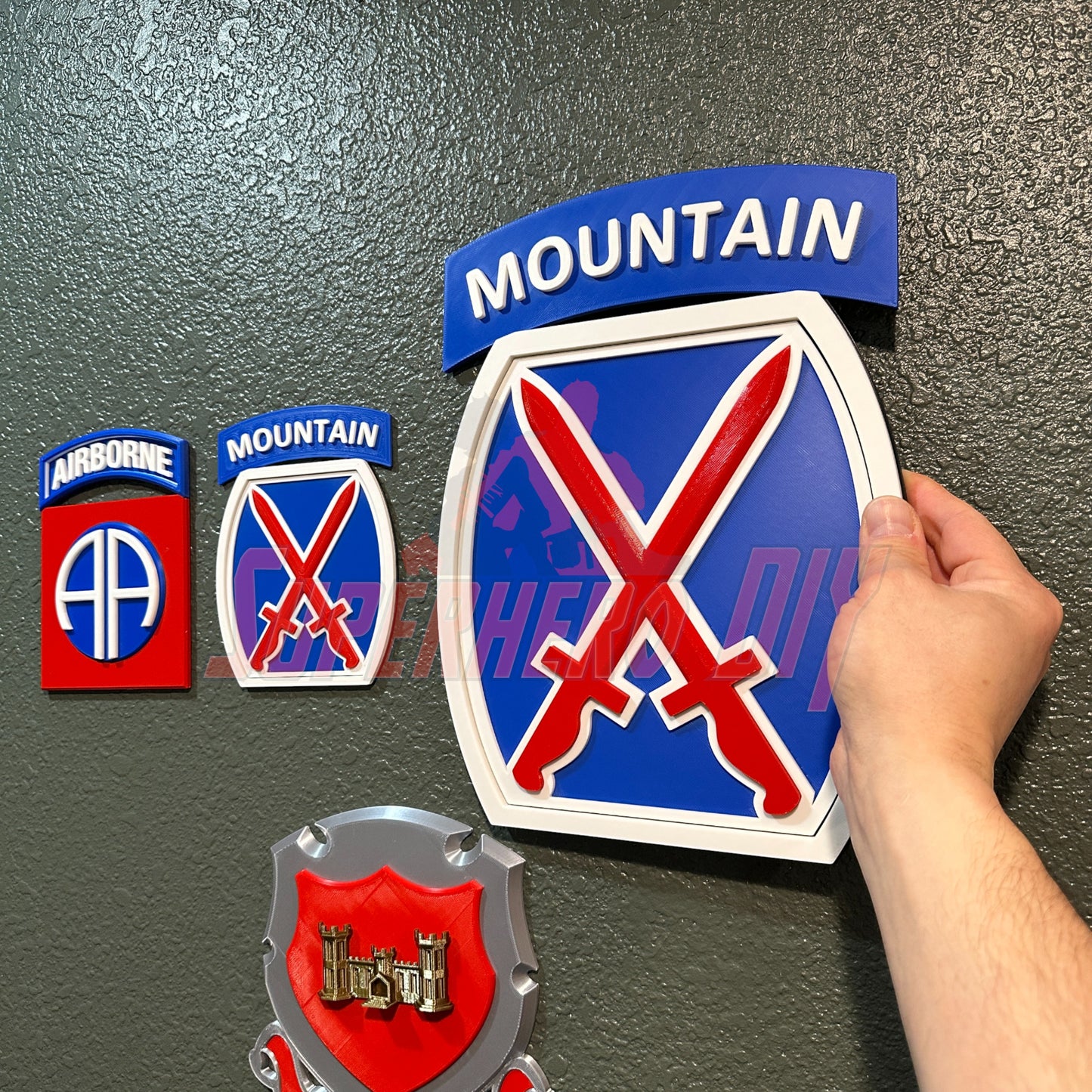 Person holding 3D-printed 10th Mountain Division insignia in blue, red, and white, alongside military awards and symbols.