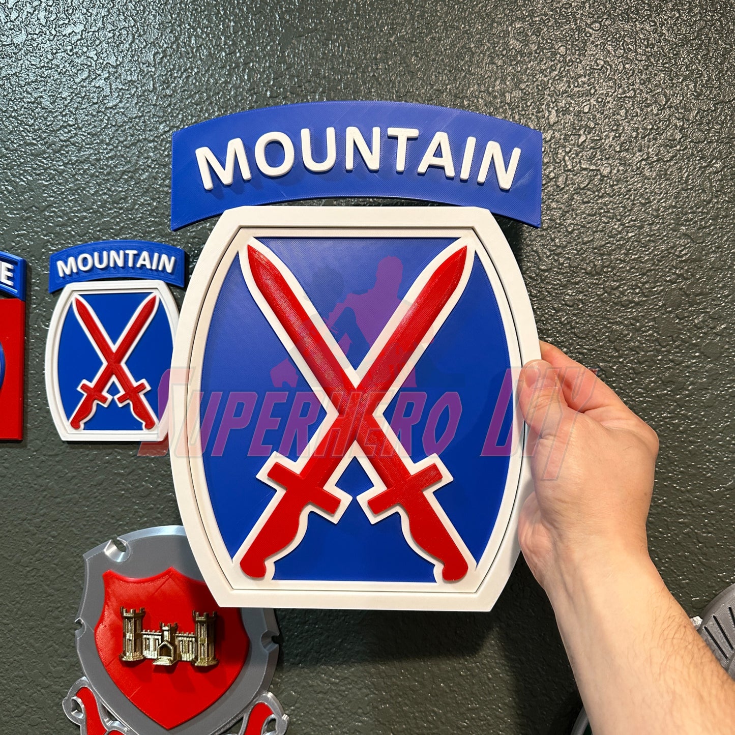 3D-Printed 10th Mountain Division insignia in blue, red, and white held by a hand, showcasing bold lettering and crossed swords design.