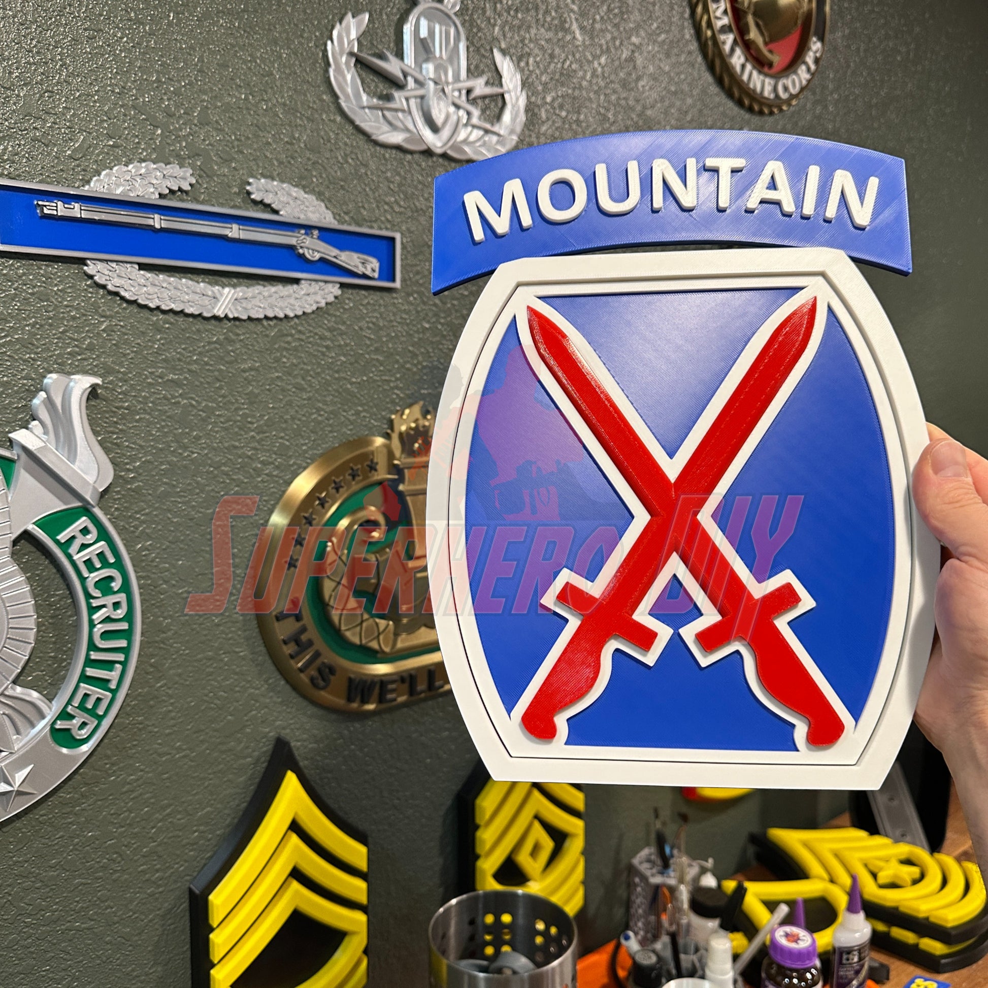 3D-printed 10th Mountain Division insignia in blue, red, and white held against a wall with military memorabilia, showcasing bold design.