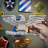Hand holding a detailed Colonel Captain rank insignia O-6 eagle emblem against a background of military patches.