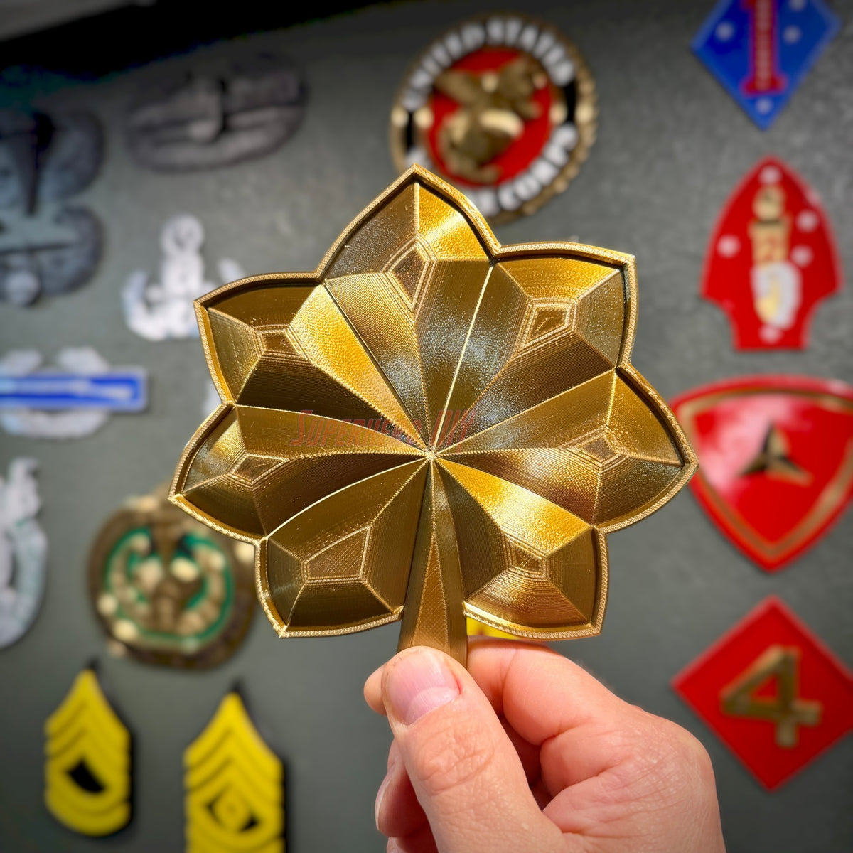 Hand holding a Major O-4 Lieutenant Commander rank insignia replica against a display of various military emblems.