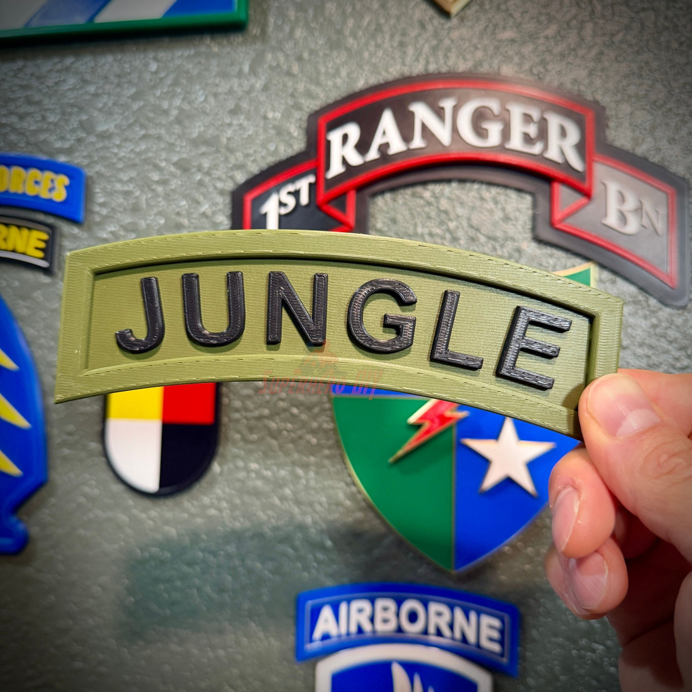 Hand holding a 3D Jungle Tab display piece with army green background and black JUNGLE lettering.
