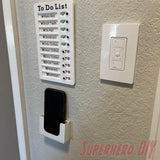 Wall-mounted phone holder under a to-do list on a bathroom wall, installed with Command strips for easy mounting.