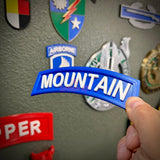 3D mountain tab wall display in hand against a backdrop of various military patches
