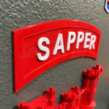 3D sapper tab display with bold red lettering on a gray surface, representing combat engineer achievement.