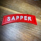 3D sapper tab displayed on a wooden surface, showcasing its bright red color and embossed white lettering.