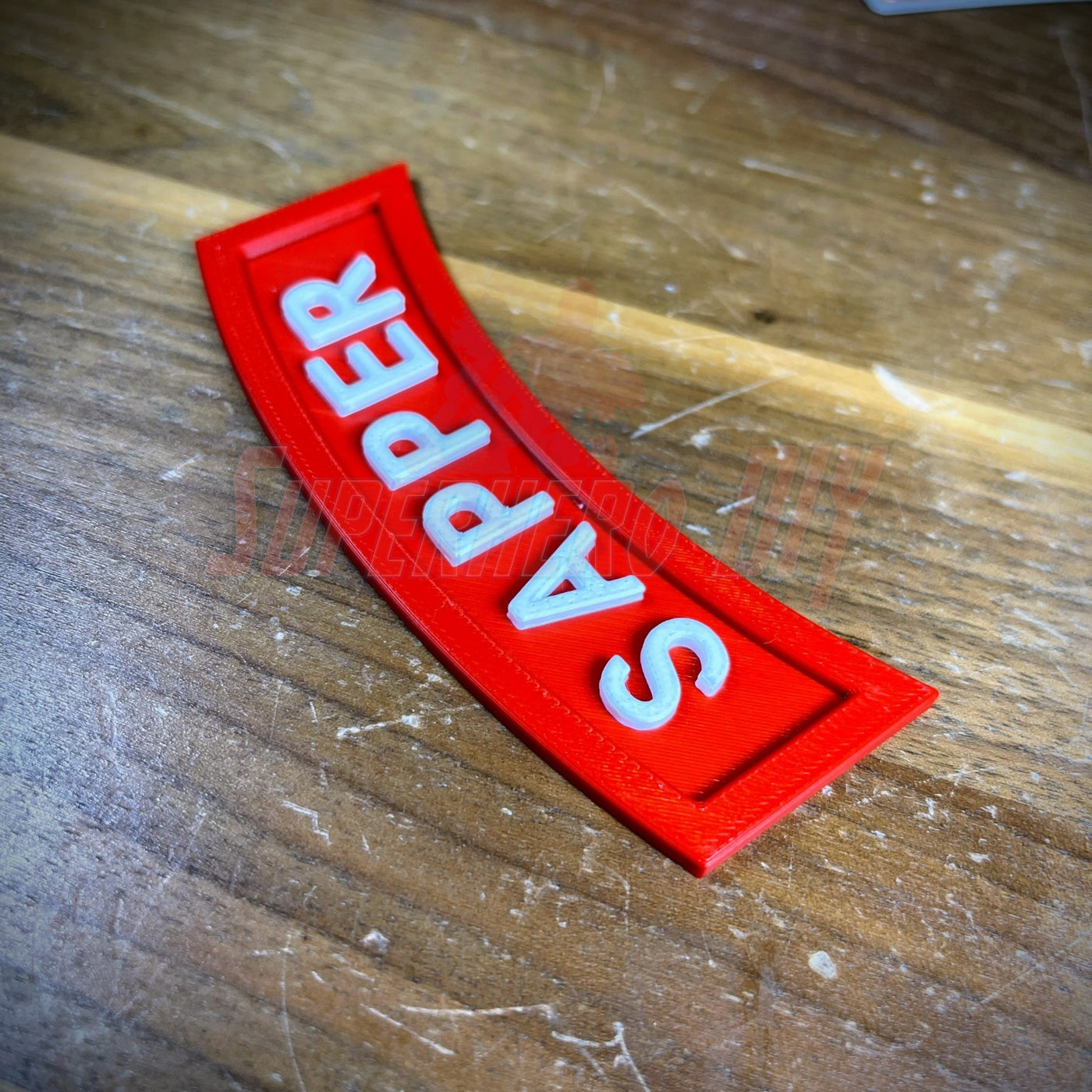3D sapper tab display piece showing red patch with white lettering on wooden surface.