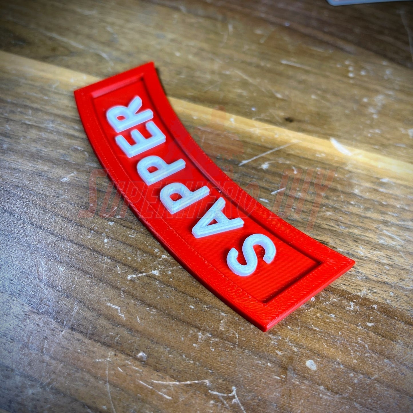 3D sapper tab display piece showing red patch with white lettering on wooden surface.