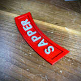 3D sapper tab display piece showing red patch with white lettering on wooden surface.