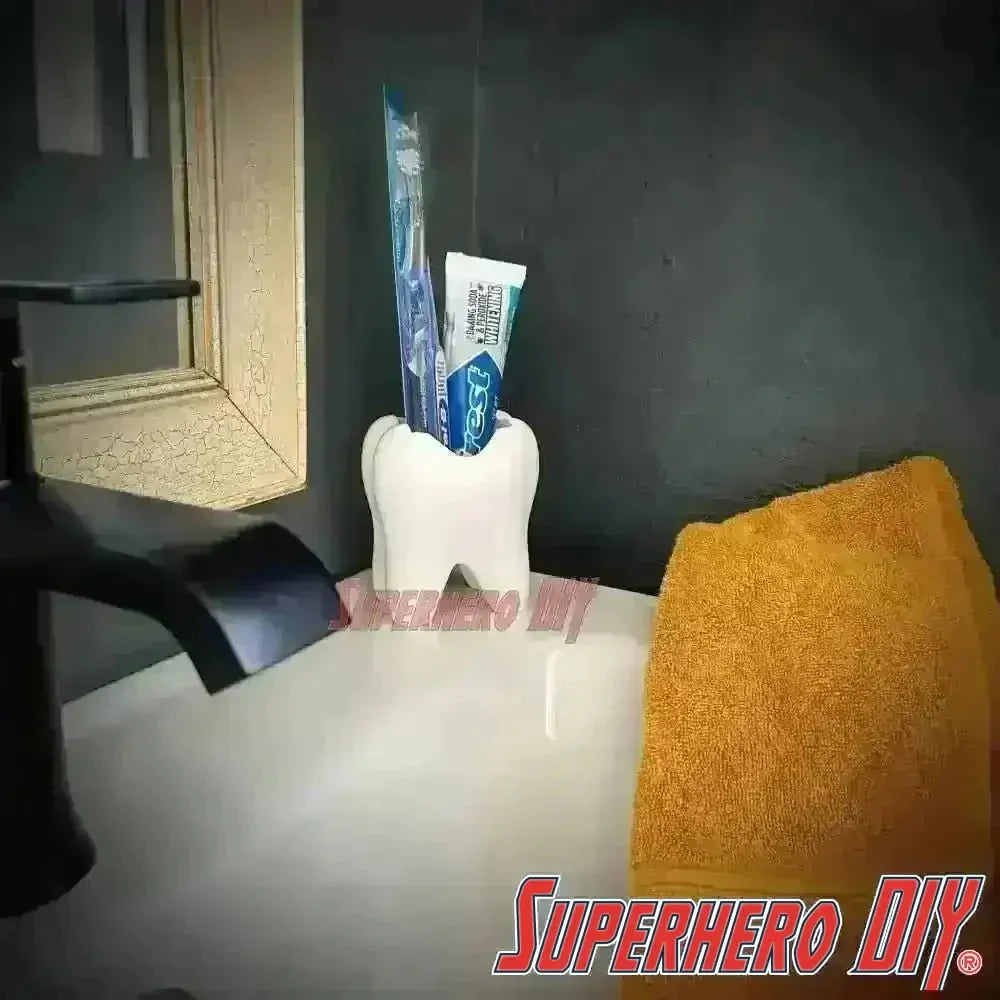 Tooth shaped bathroom organizer holding toothbrush and toothpaste placed on a modern sink with a yellow towel beside it.