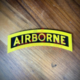3D airborne tab with bold yellow lettering on a solid black background, placed on a wooden surface.