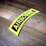 3D airborne tab displayed on a wooden surface, featuring bold yellow lettering on a black background.