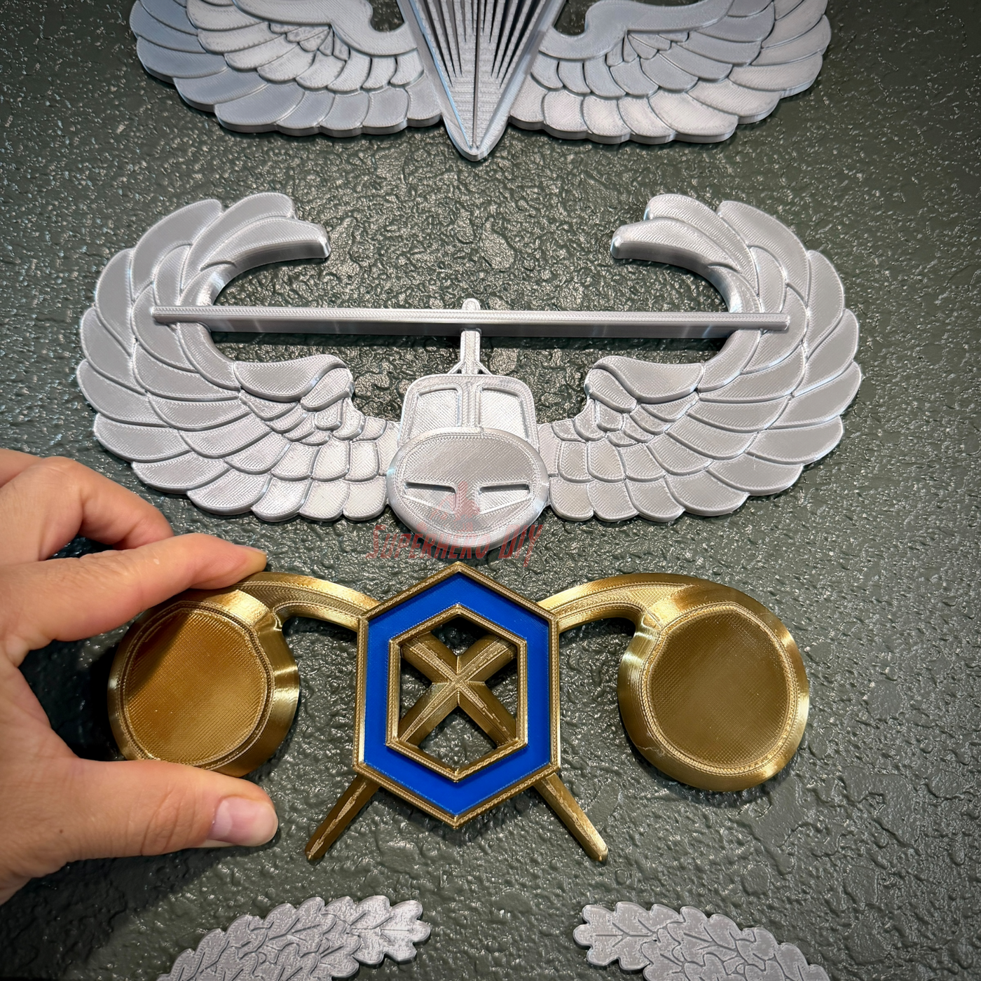Hand holding Chemical Corps insignia with wings and emblems for display on a textured wall.