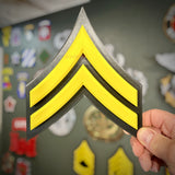 Hand holding a Corporal E-4 rank insignia, two yellow chevrons with a dark gray border, other insignia blurred behind
