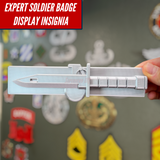 Expert Soldier Badge display insignia shown in silver on a wall with various military patches in the background.