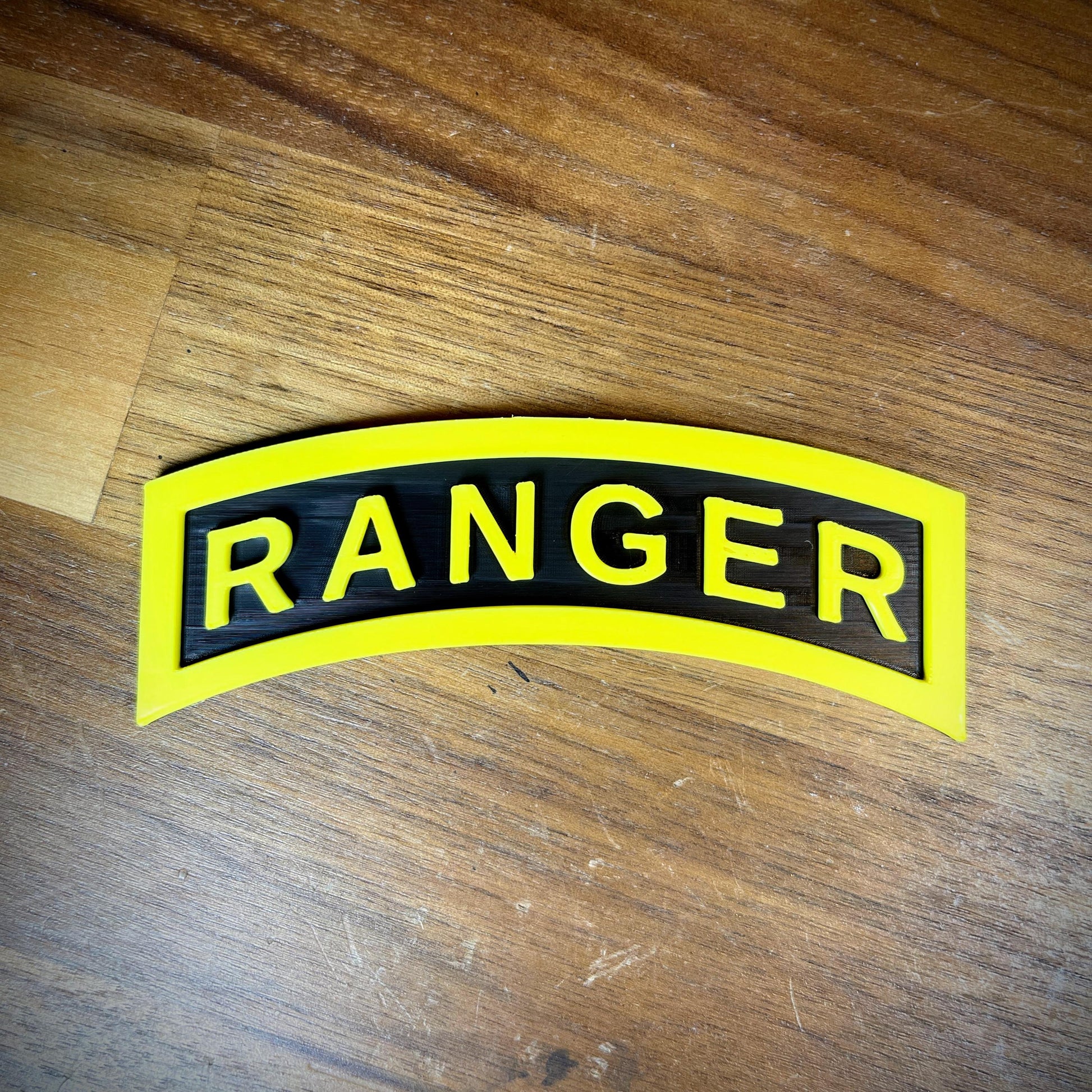 3D Army Ranger Tab replica displayed on wooden surface, showcasing its iconic yellow-on-black design.