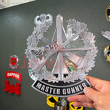 Master Gunner Badge display insignia held by hand in front of wall decor.