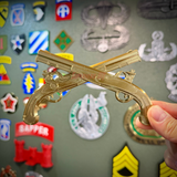 Army military police insignia, crossed pistols display in gold held against a background of military patches.
