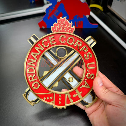 Ordnance regimental crest displayed in hand, featuring vibrant red and gold colors, symbolizing the Ordnance Corps.