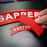 3D sapper tab display piece showing red SAPPER tab with a hand holding a removable piece.