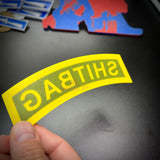 A person holding a SHITBAG tab in classic arched style, showcasing yellow and green colors for a gag gift.
