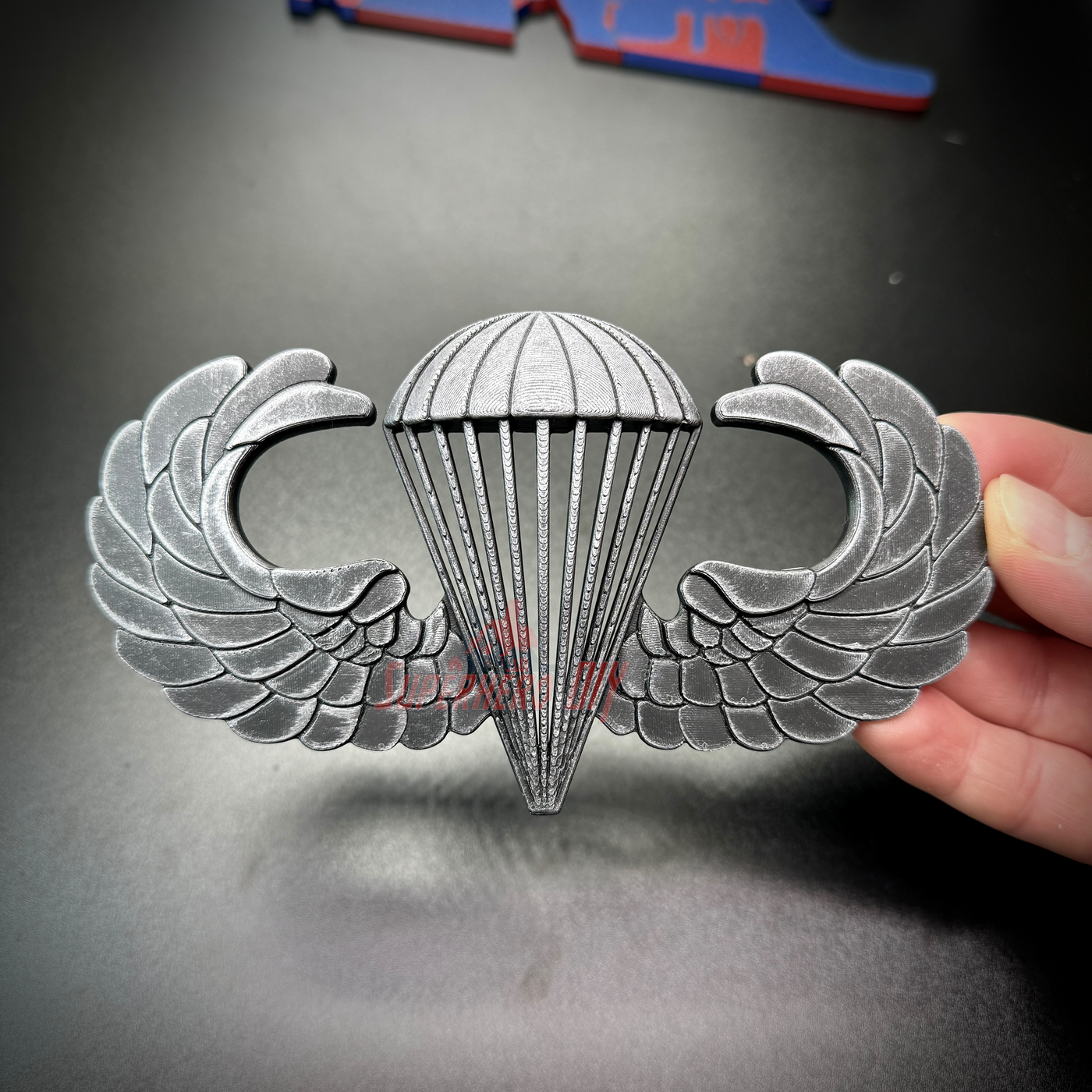 Subdued airborne jump wings replica badge, showcasing detailed design of parachute and wings.