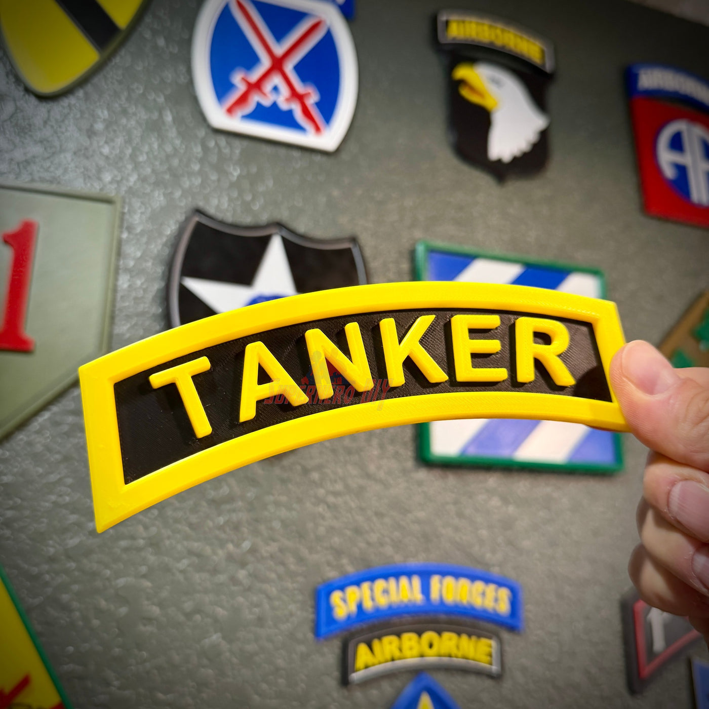 3D tanker tab held in hand with yellow raised lettering displayed among military patches on a textured wall.