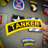 3D tanker tab held in hand with yellow raised lettering displayed among military patches on a textured wall.