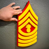 A large USMC enlisted rank insignia for Master Gunnery Sergeant displayed on the wall with hand for scale, featuring classic red and yellow design.